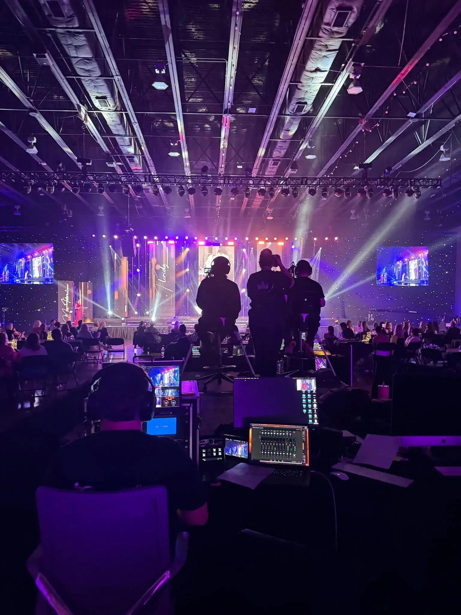 Large purple-lit event with stage, audience, cameras, consoles, and production crew
