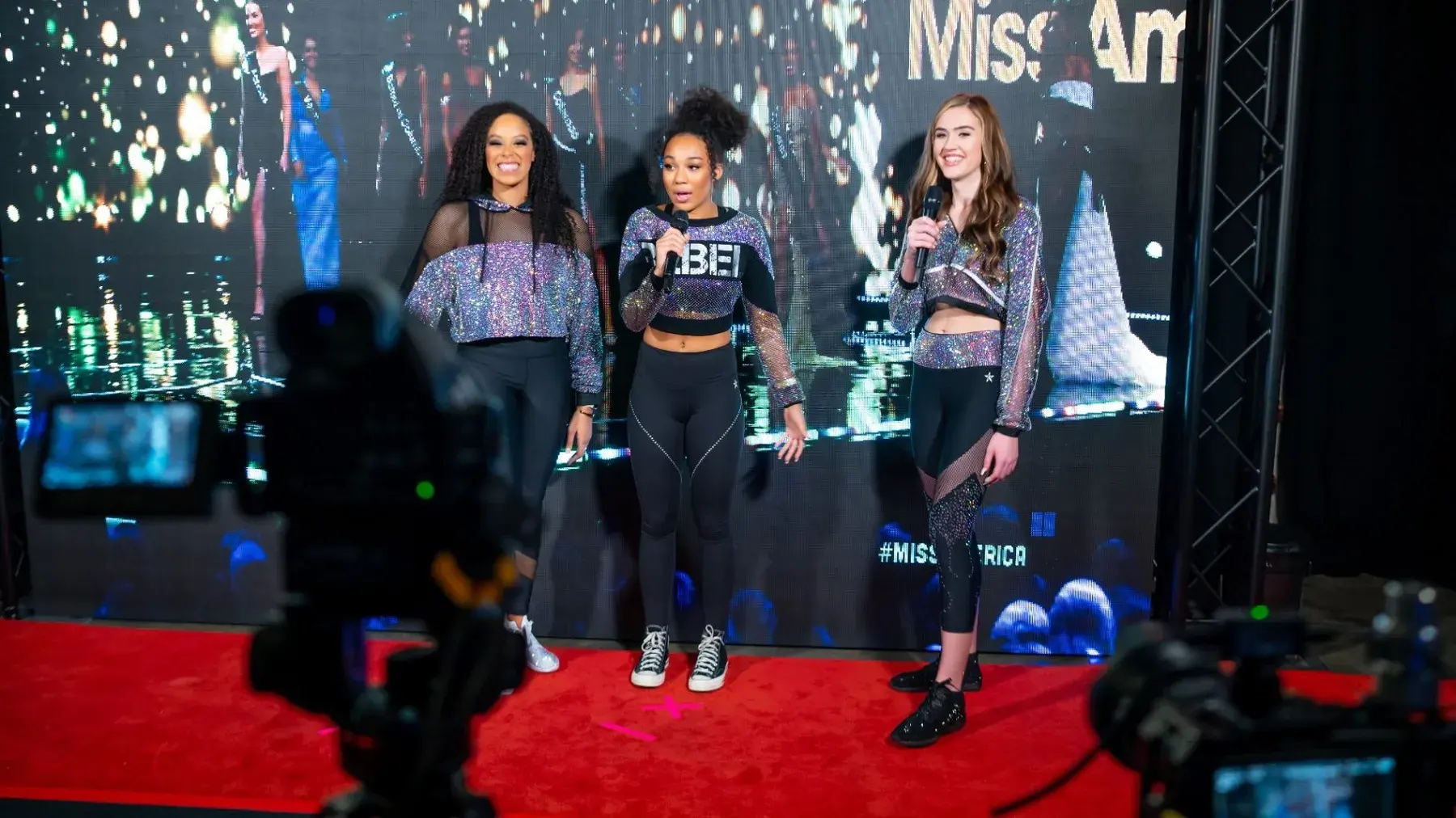 Presenters speaking on stage with cameras and a video wall behind them