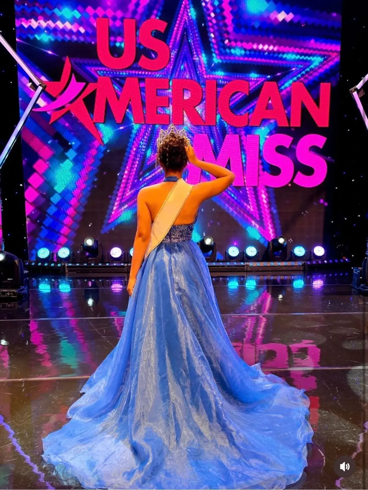 US American Miss contestant in a blue gown looking toward the colorful pageant logo on a video wall